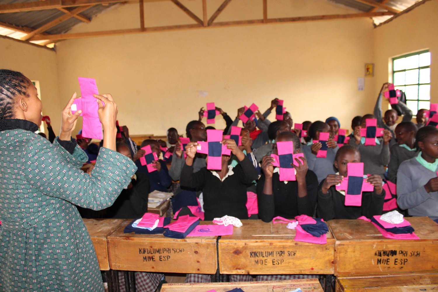 Students holding up pink and navy reusable sanitary pads in a classroom at Kikulumi Primary School, led by a female teacher.