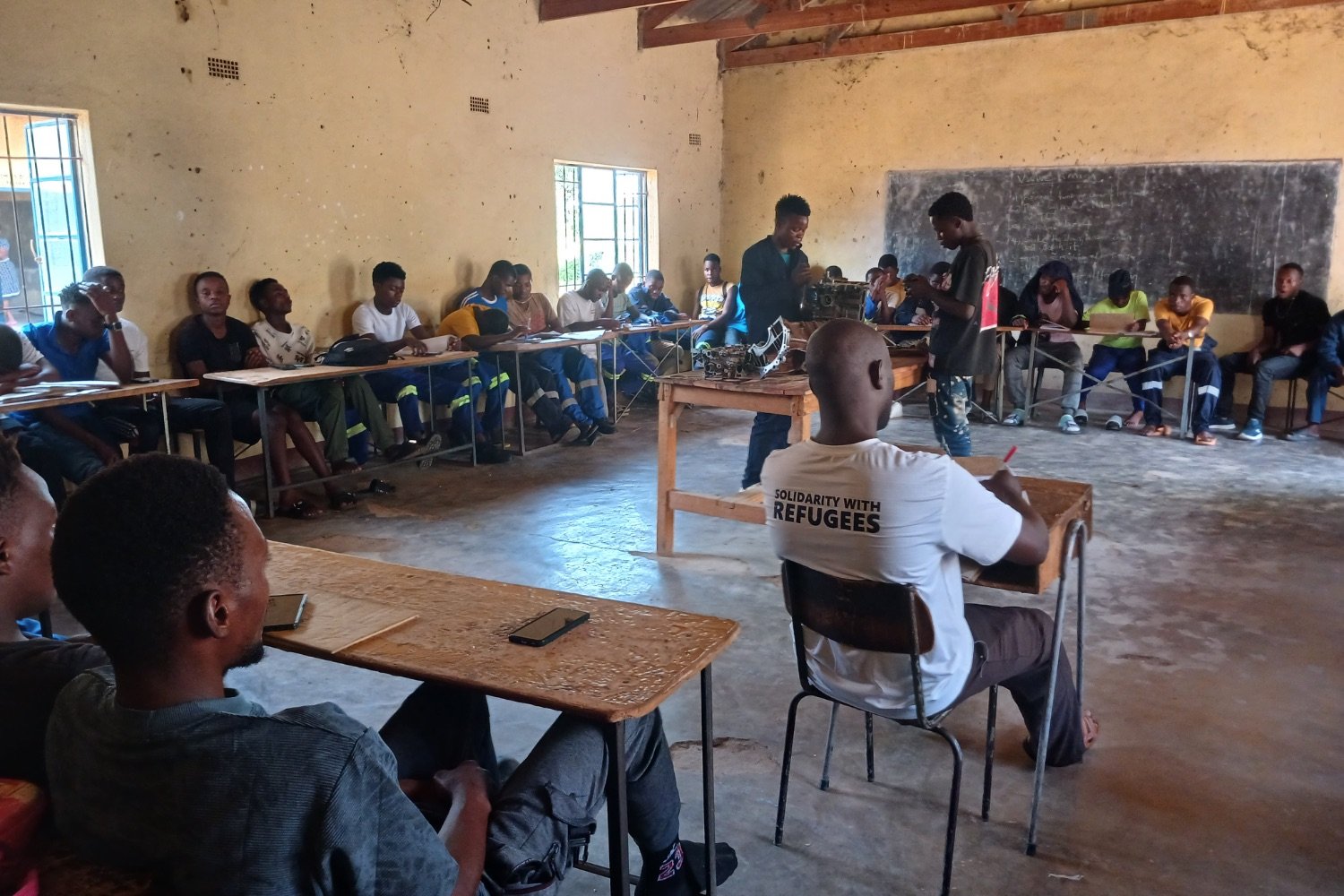 Students sitting at wooden desks in a basic classroom while two peers present something at the front near a chalkboard.
