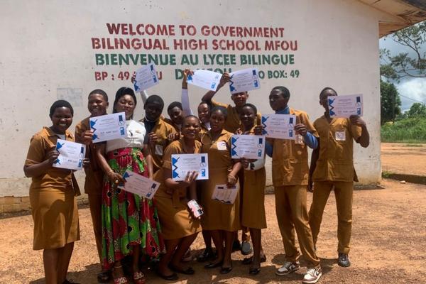 Scholars in brown uniforms hold their graduation certificates in front of the Government Bilingual High School Mfou sign.
