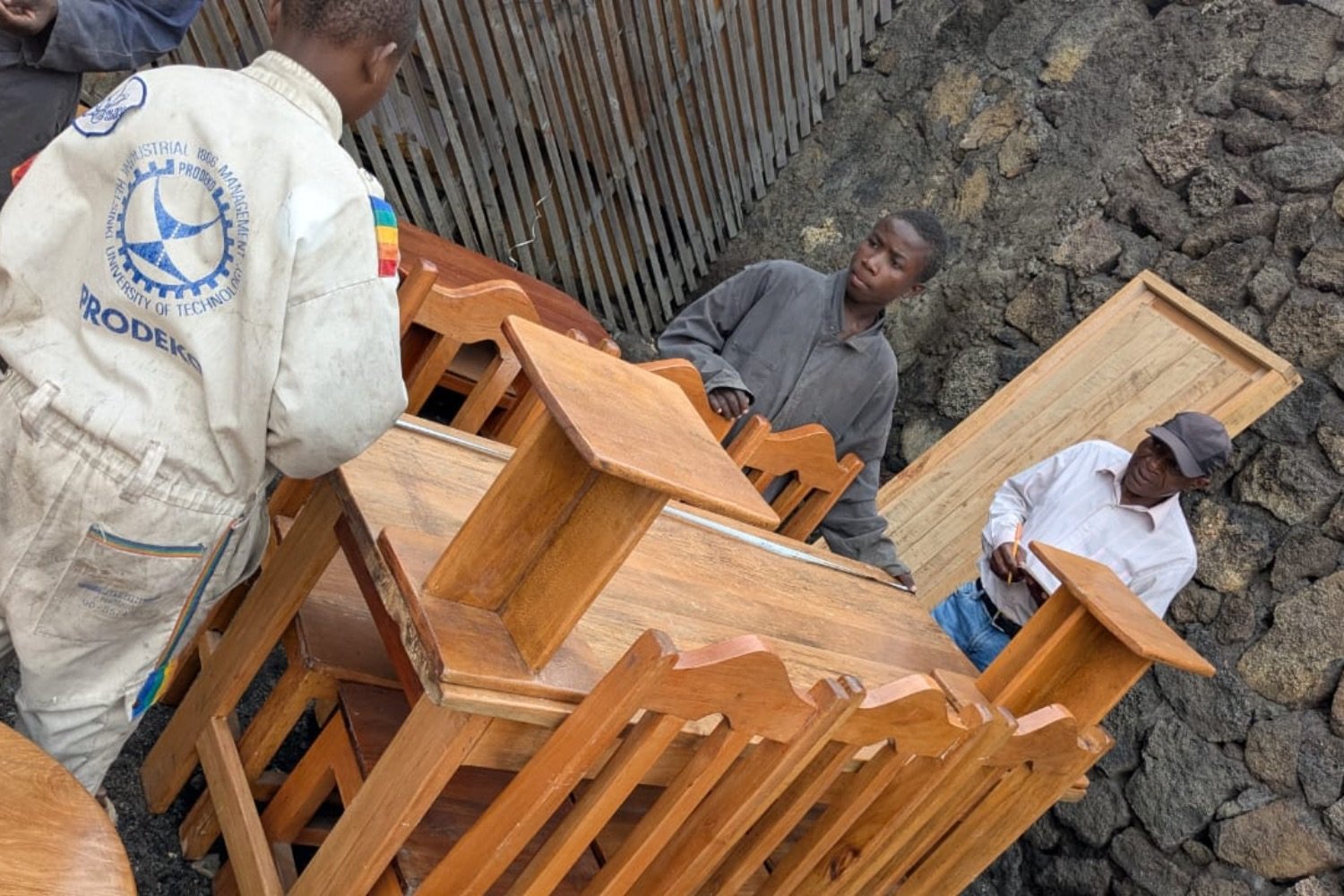 Workers unloading wooden school desks and chairs onto rocky ground near a corrugated metal fence.