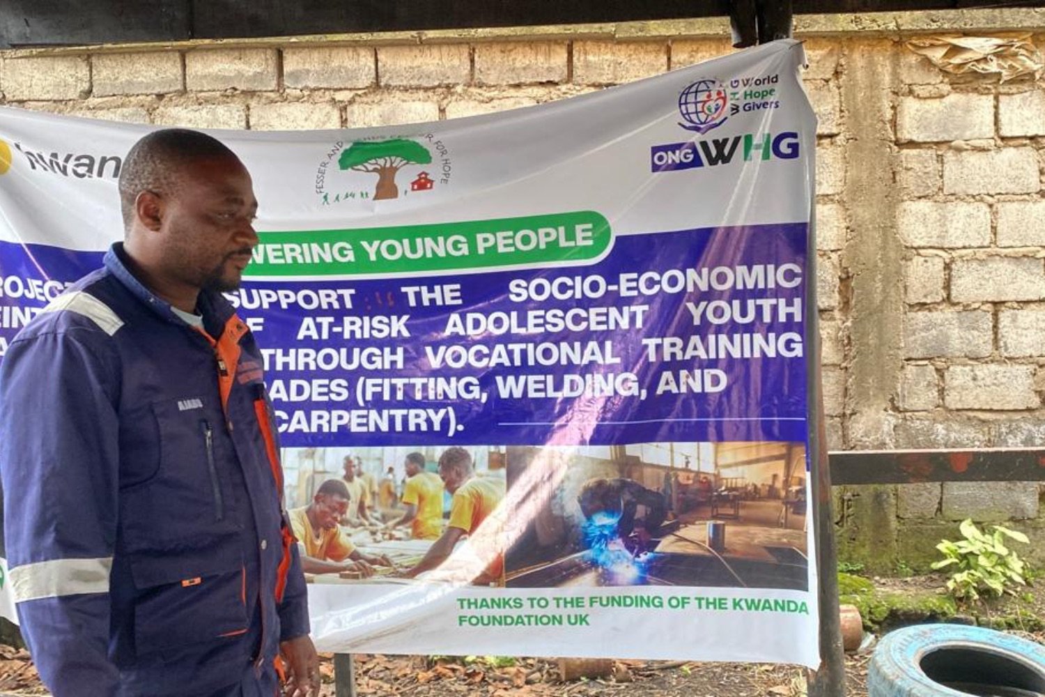 Man in blue coveralls standing beside a banner for ONG WHG's vocational training program for at-risk youth in an outdoor setting.