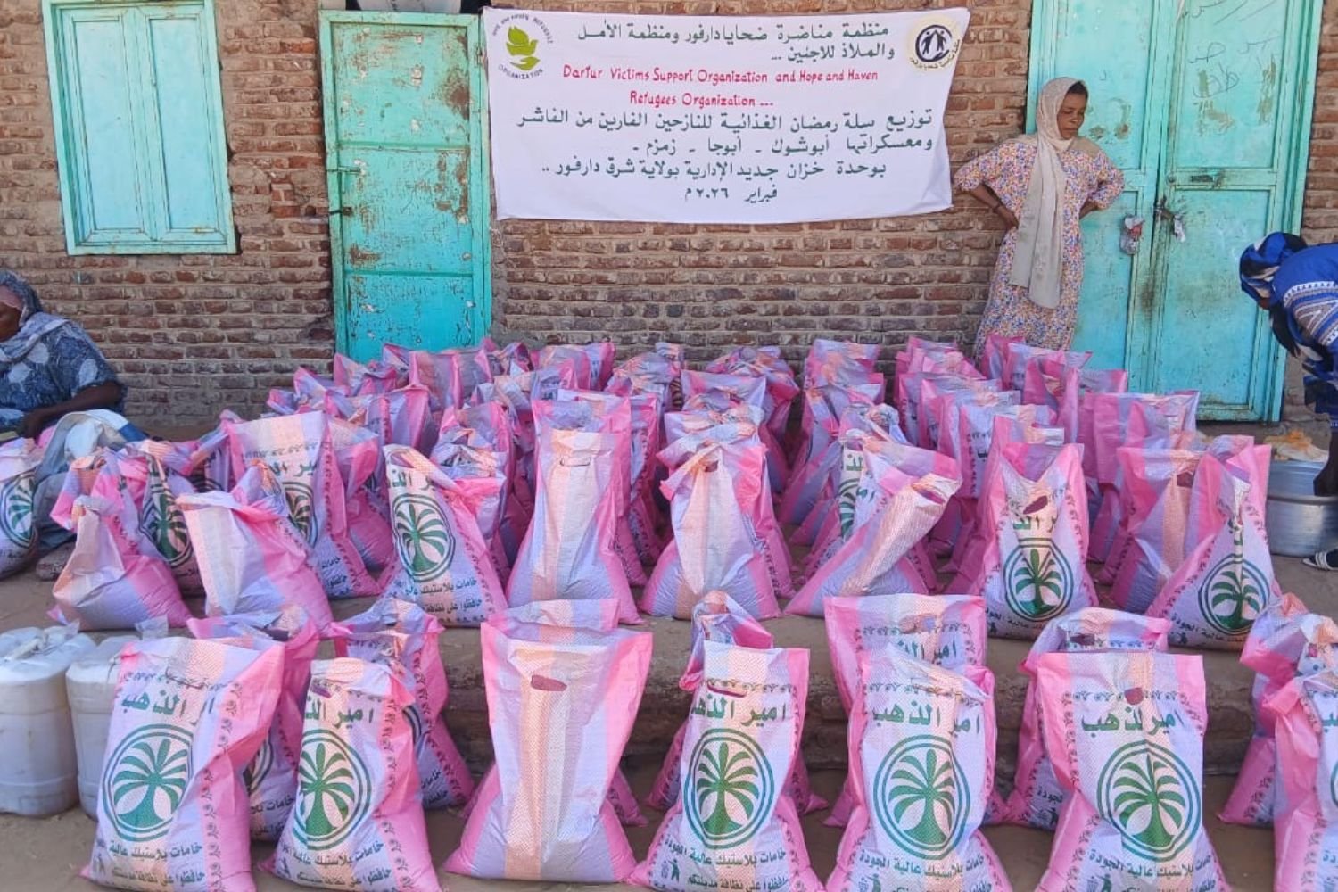 Rows of pink and white branded food sacks arranged outside a brick building beneath an Arabic and English banner in Darfur.