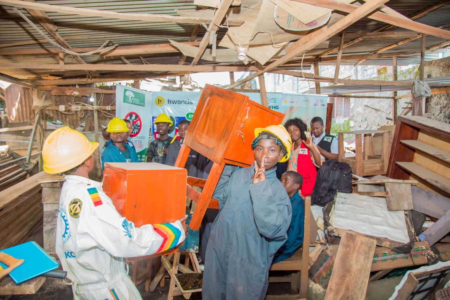 Workers in yellow hard hats removing orange furniture from a damaged wooden structure with a partially collapsed roof.
