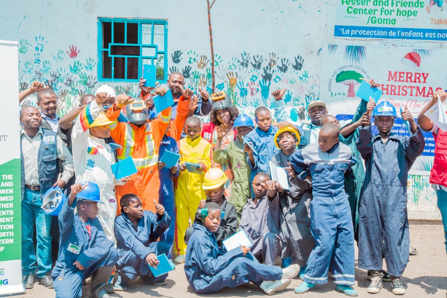 Children and adults in work uniforms and hard hats holding notebooks outside the Fesser and Friends Center for Hope in Goma.