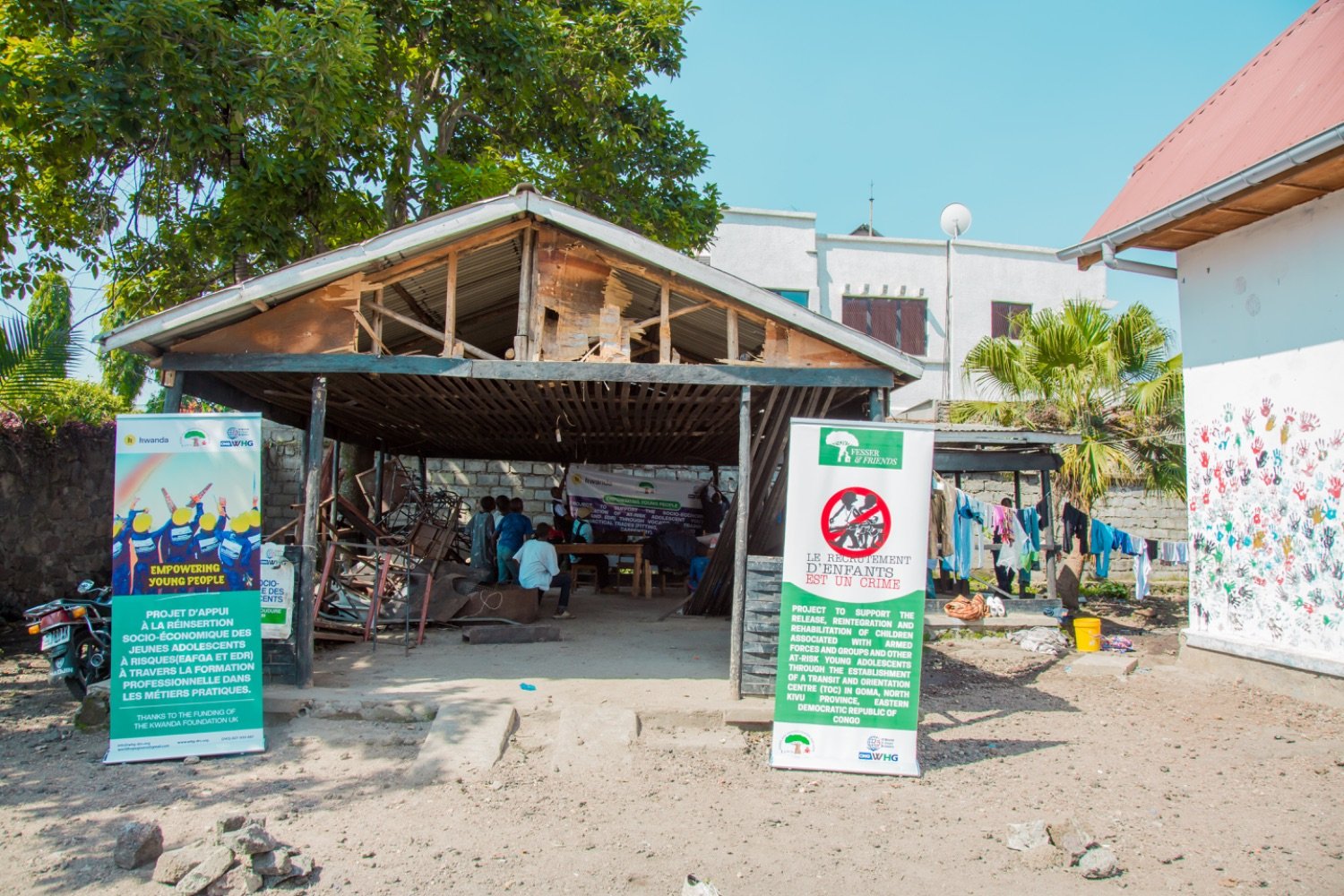 People attending a meeting under an open-air shelter flanked by NGO banners about child soldier rehabilitation in Goma, Democratic Republic of Congo.