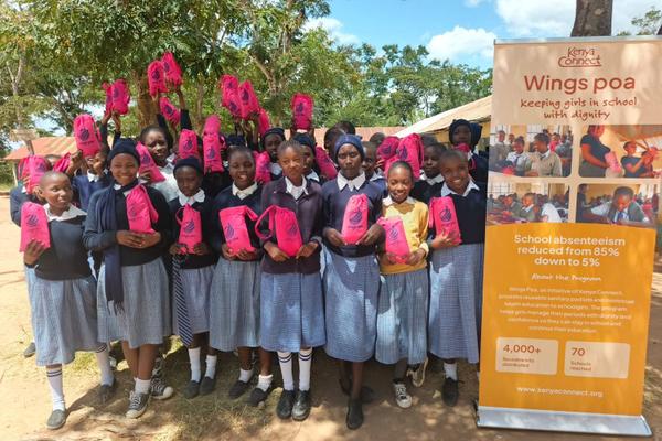 Schoolgirls in blue uniforms hold pink drawstring bags outdoors beside a WingsPoa banner.