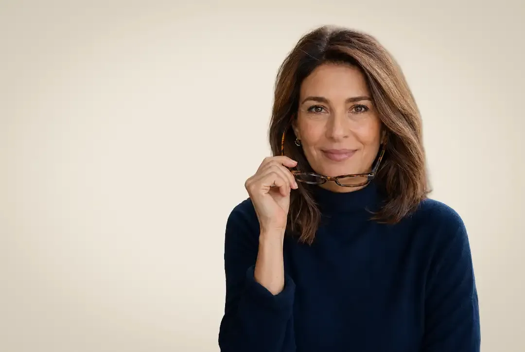 Frau mit braunen Haaren in einem marineblauen Rollkragenpullover, die eine Lesebrille in der Hand hält und vor einem beigen Hintergrund lächelt.