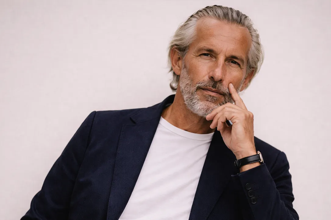 Image Mann mit grauen Haaren und Bart in einem marineblauen Blazer und einem weißen T-Shirt, der nachdenklich mit der Hand am Kinn vor einem hellen Hintergrund sitzt.