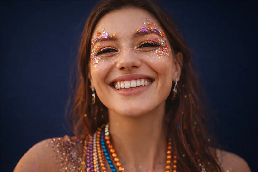 Lächelnde Frau mit buntem Glitzer-Make-up und Perlenketten vor einem dunkelblauen Hintergrund.