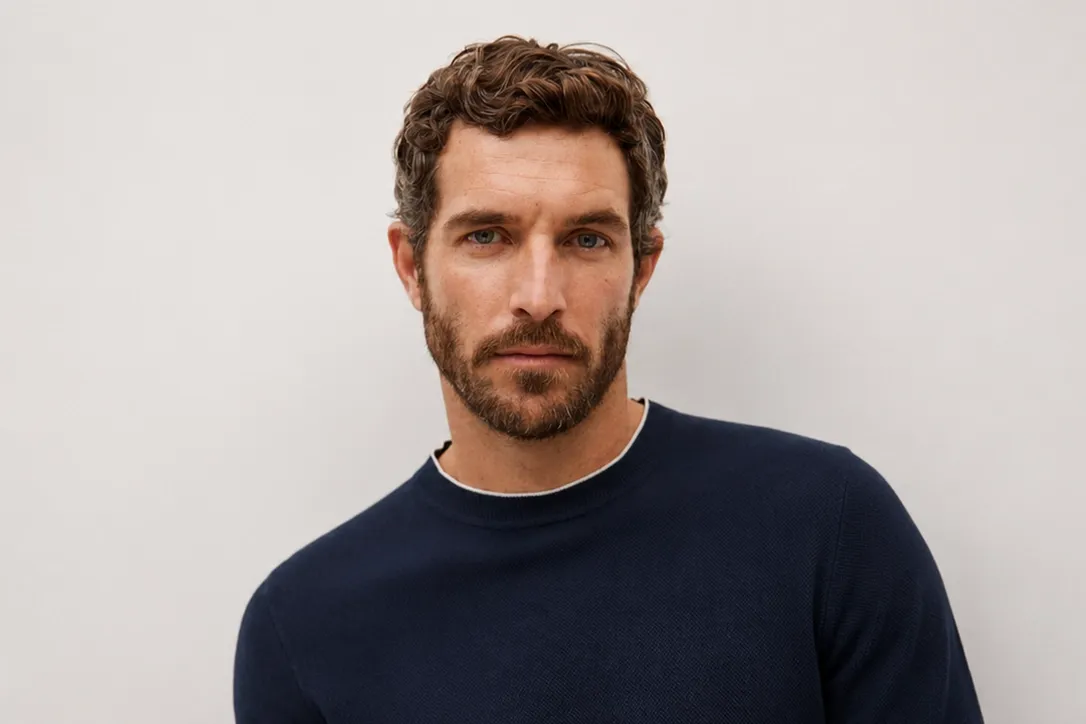 Ein Mann mit lockigem braunem Haar und Bart, der einen marineblauen Pullover mit Rundhalsausschnitt trägt und vor einem hellgrauen Hintergrund posiert.