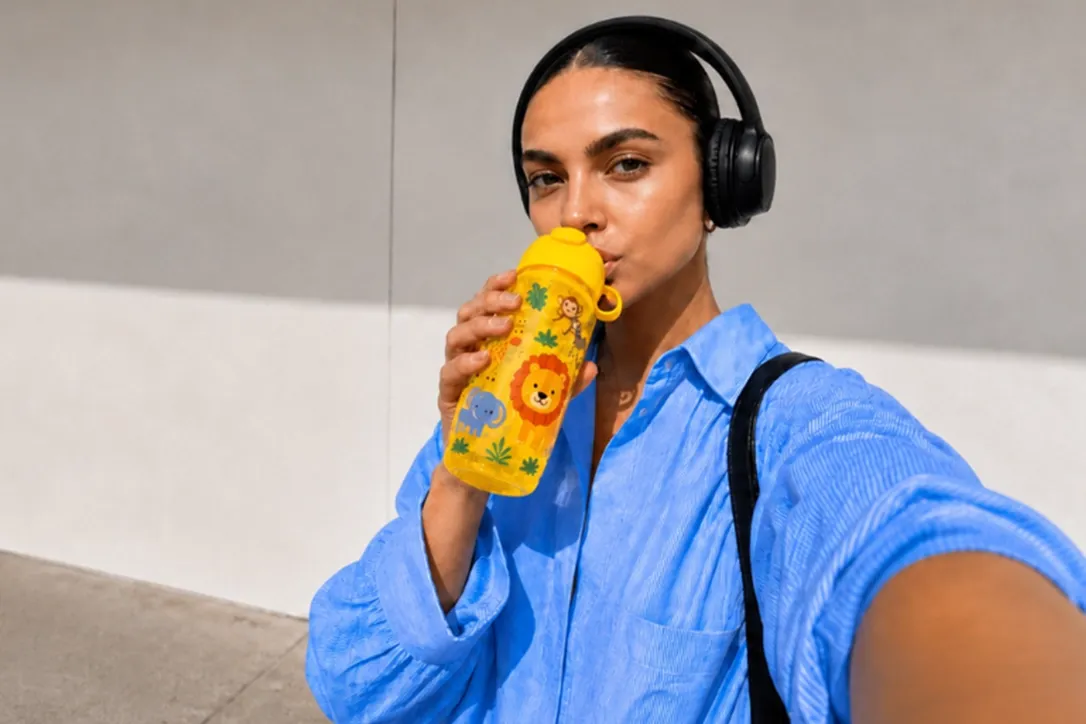 Eine Frau in einem blauen Shirt und mit schwarzen Kopfhörern trinkt aus einer gelben Kinder-Trinkflasche mit Dschungeltiermotiven.