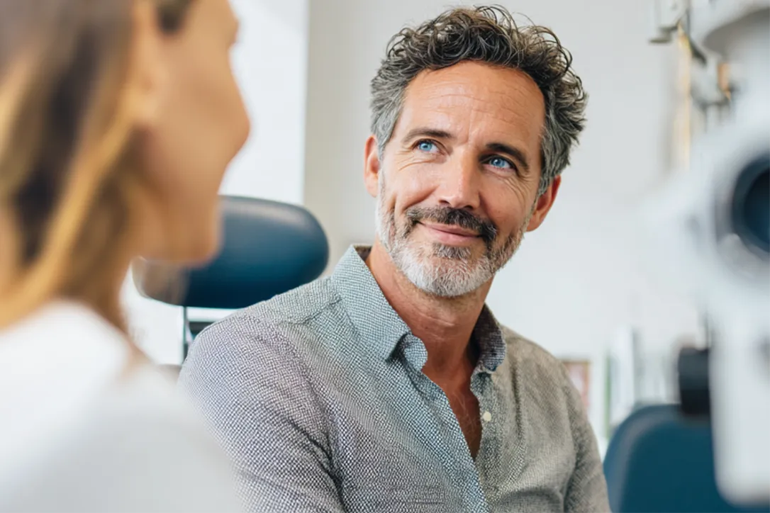 Ein Mann der bei einer Augenvoruntersuchung im Behandlungszimmer sitz und zur Augenärztin schaut.