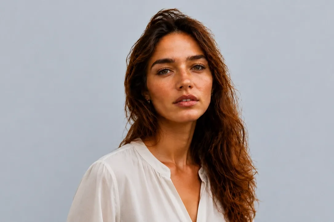 Image Junge Frau mit langen, rotbraunen, gewellten Haaren und Sommersprossen, die eine weiße Bluse trägt und vor einem grauen Hintergrund in die Kamera blickt.
