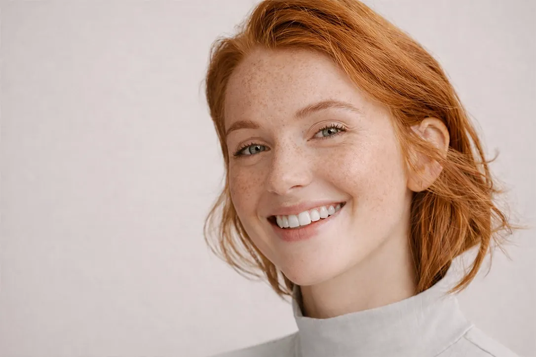 Image Lächelnde Person mit roten Haaren, Sommersprossen und einer klaren Brille, die einen hellgrauen Rollkragenpullover vor einem einfarbigen Hintergrund trägt.