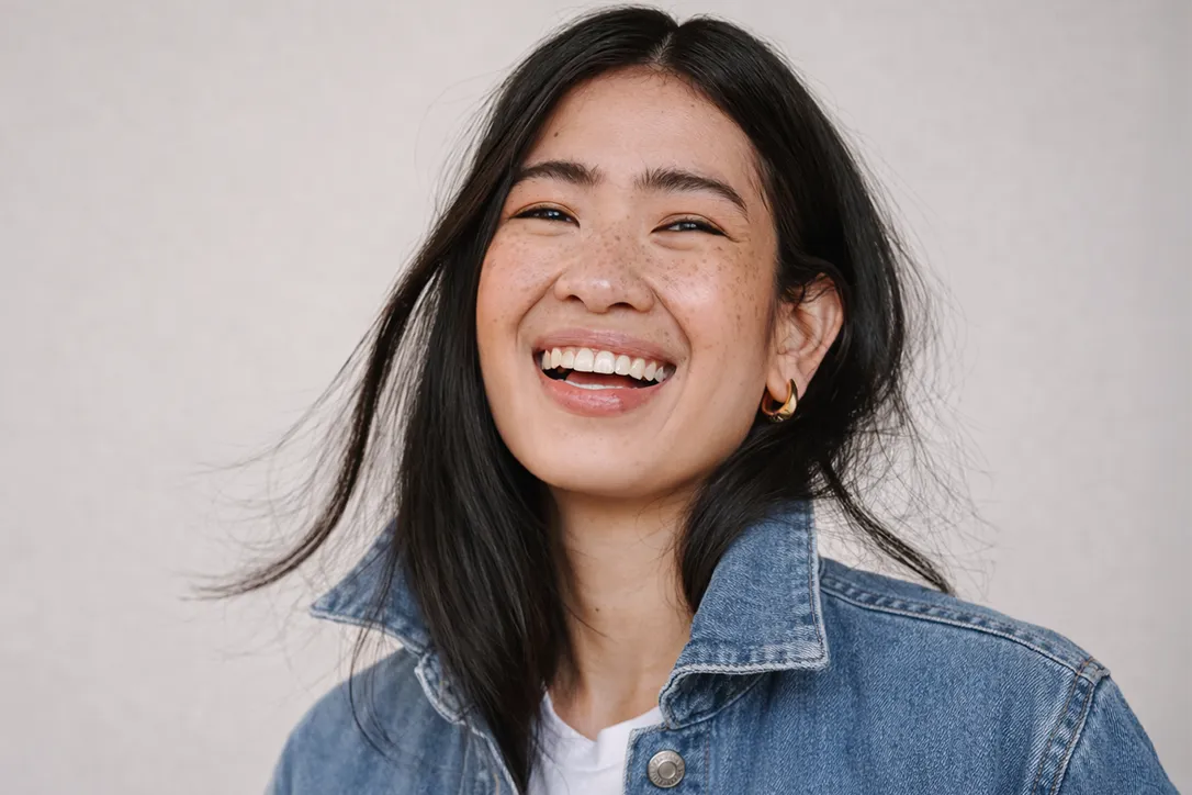 Image Lächelnde Person mit langen dunklen Haaren, die eine Jeansjacke und goldene Creolen trägt, vor einem einfarbigen Hintergrund.