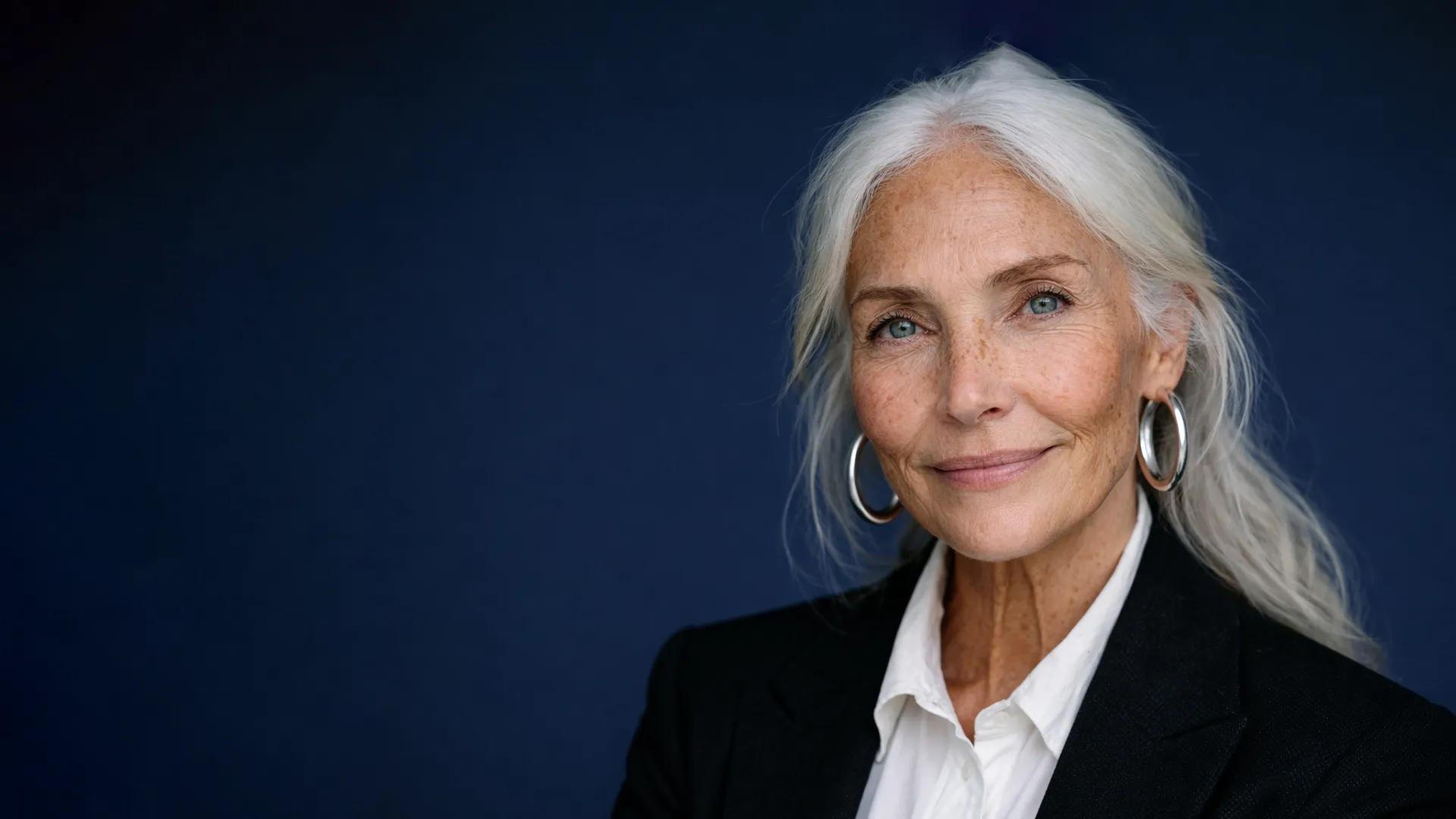 Eine elegante ältere Frau mit langem silbernem Haar, blauen Augen, Creolen und einem schwarzen Blazer, die vor einem dunkelblauen Hintergrund lächelt.