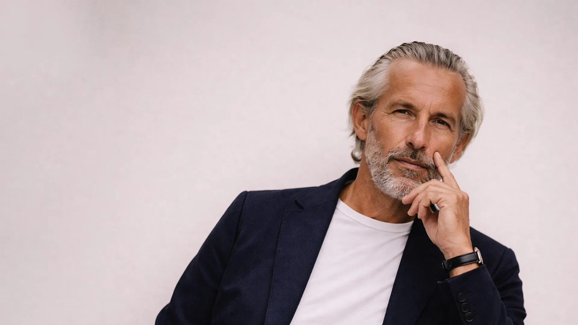 Mann mit grauen Haaren und Bart in einem marineblauen Blazer und einem weißen T-Shirt, der nachdenklich mit der Hand am Kinn vor einem hellen Hintergrund sitzt.