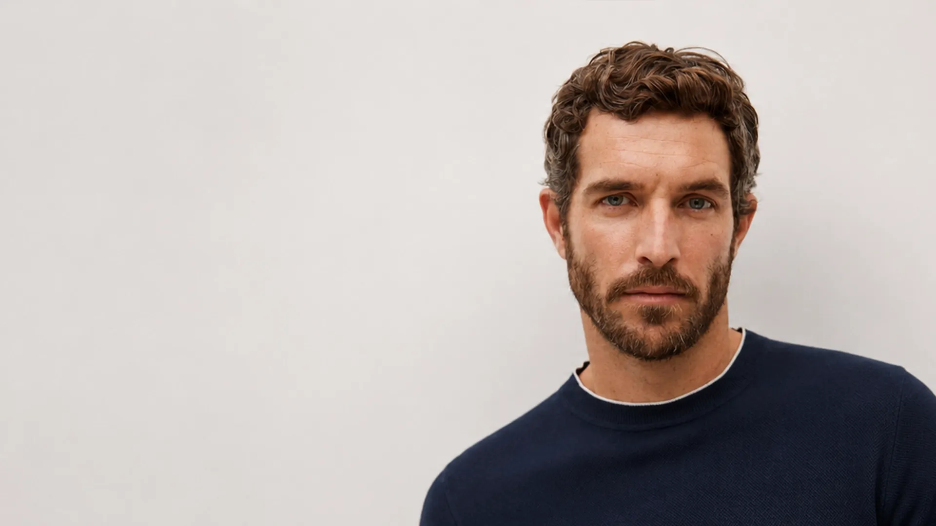 Ein Mann mit lockigem braunem Haar und Bart, der einen marineblauen Pullover mit Rundhalsausschnitt trägt und vor einem hellgrauen Hintergrund posiert.