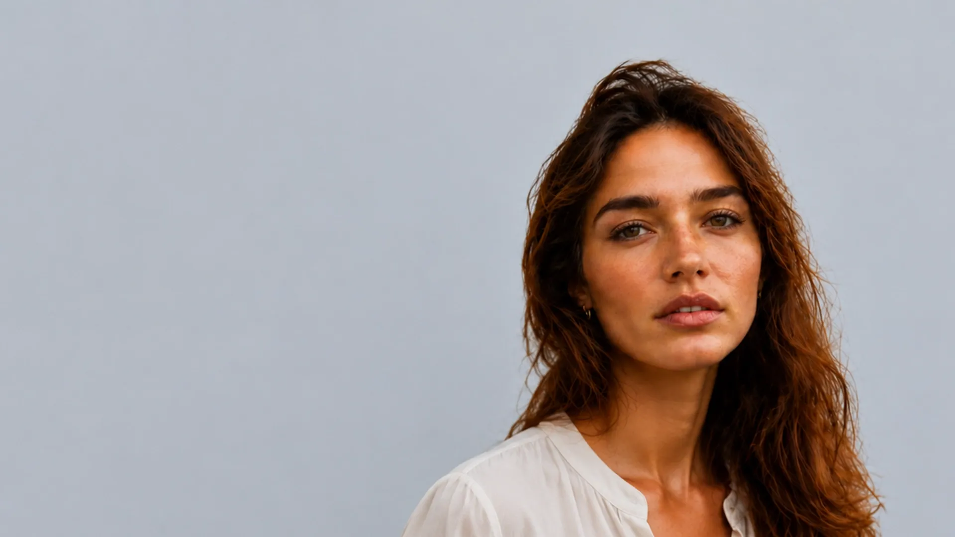 Junge Frau mit langen, rotbraunen, gewellten Haaren und Sommersprossen, die eine weiße Bluse trägt und vor einem grauen Hintergrund in die Kamera blickt.