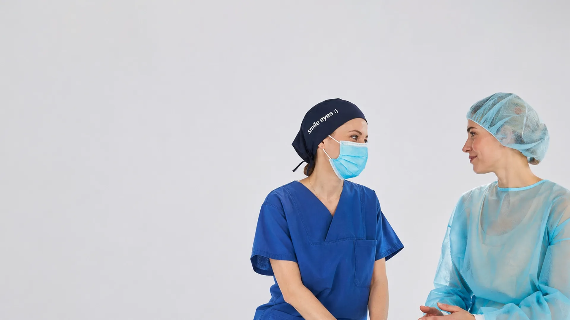 Zwei medizinische Fachkräfte in OP-Kleidung und OP-Hauben unterhalten sich; eine trägt eine blaue Maske und eine Mütze mit „Smile Eyes“, die andere einen hellblauen Kittel.