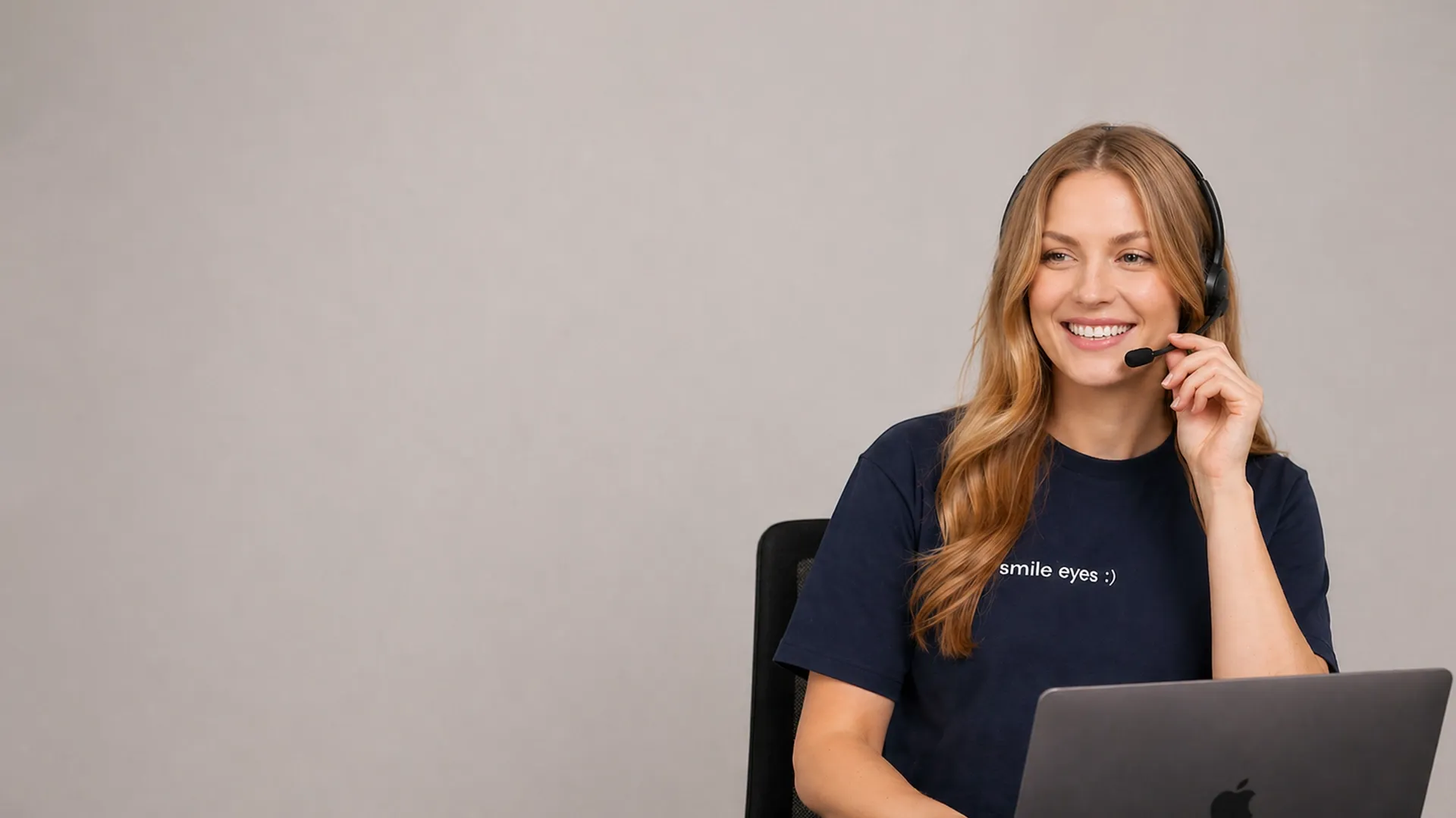 Eine lächelnde Kundendienstmitarbeiterin mit Headset und einem T-Shirt mit der Aufschrift „Smile Eyes“, die vor einem grauen Hintergrund an einem Laptop sitzt.