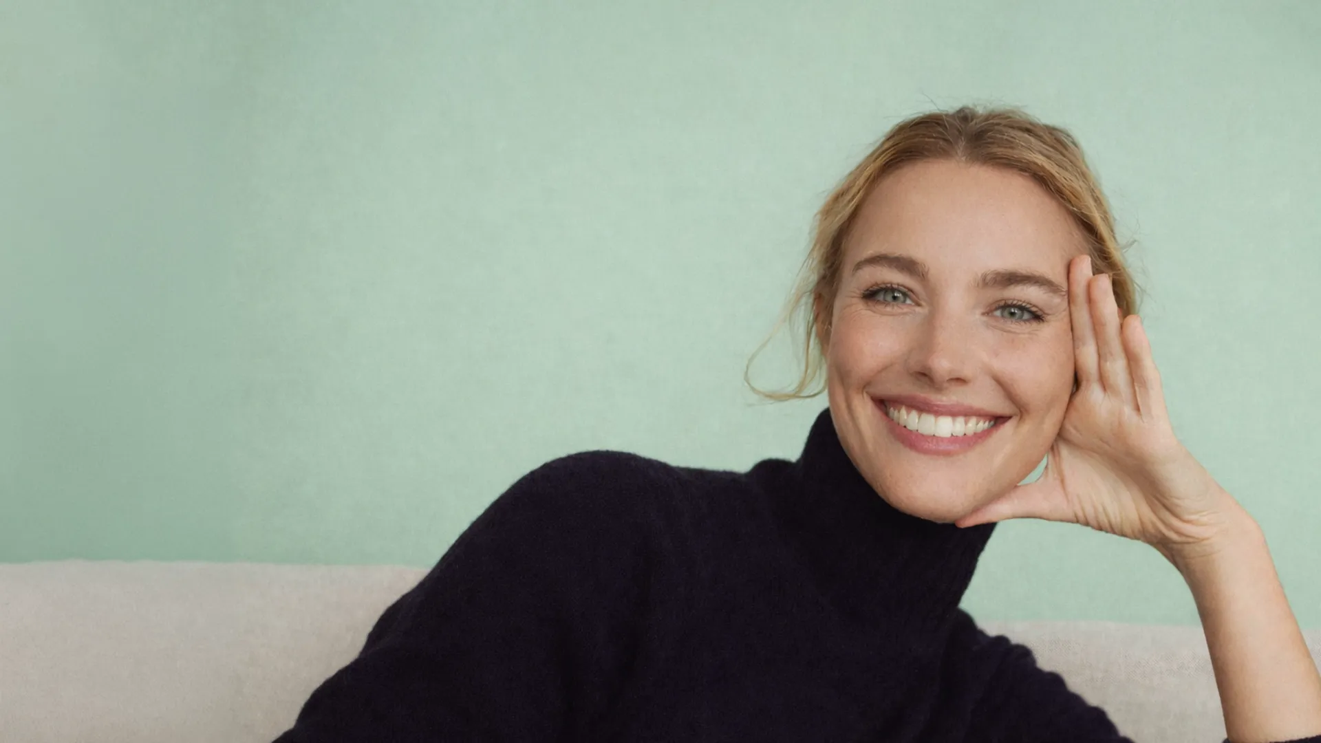 Eine lächelnde blonde Frau in einem schwarzen Rollkragenpullover, die eine Hand an die Wange legt, vor einem zart mintgrünen Hintergrund.