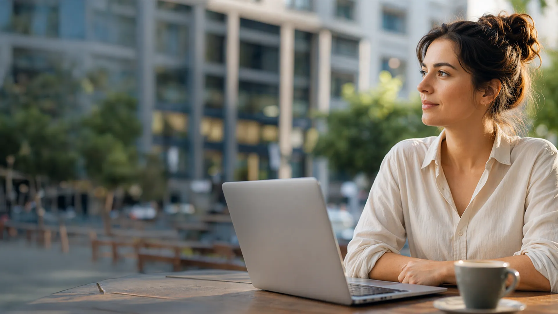 Eine Frau in einem weißen Hemd sitzt draußen an einem Tisch, blickt zur Seite und hat einen Laptop und eine Kaffeetasse vor sich stehen. Städtischer Hintergrund.