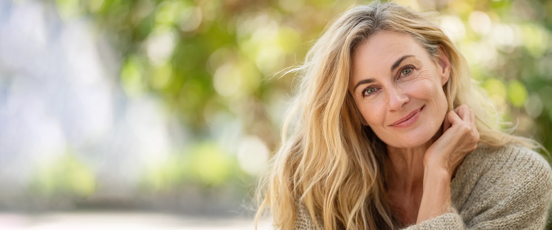Eine Frau mit langen blonden Haaren lächelt warm, ihr Kinn auf die Hand gestützt, vor einem verschwommenen grünen Hintergrund.