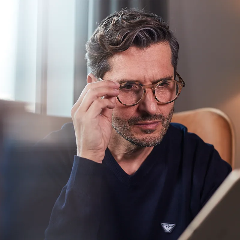 Mann mit Brille, der ein Buch liest, auf einem Stuhl sitzt, einen dunklen Pullover trägt und nachdenklich dreinschaut.