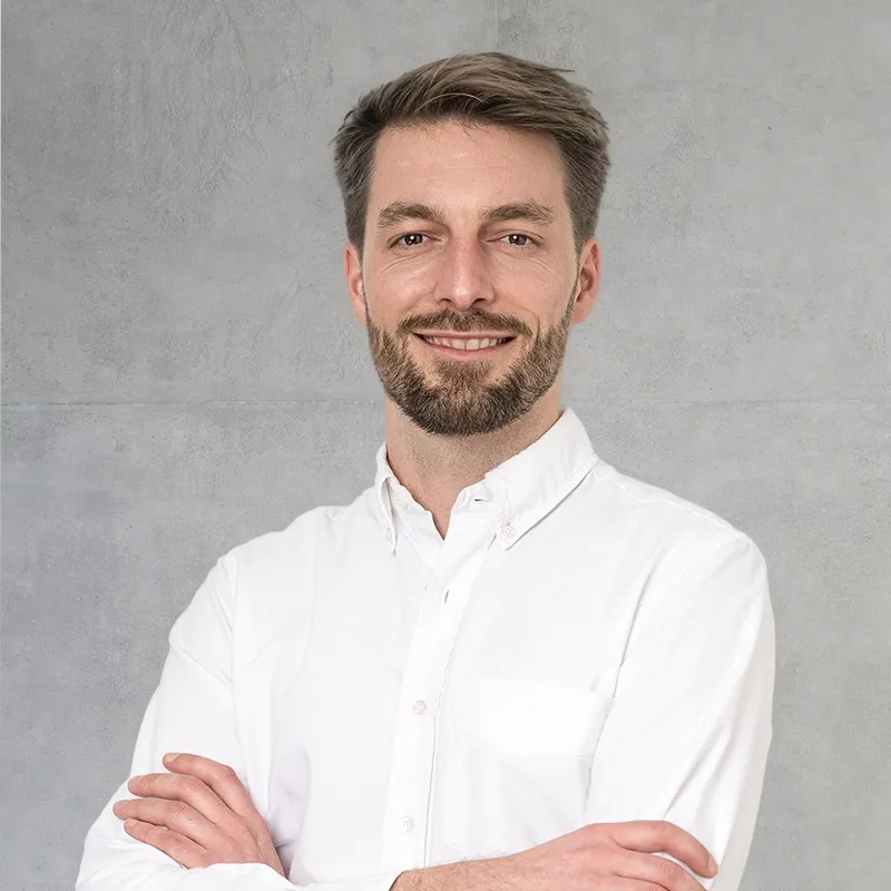 Augenarzt Klemens Waser aus Trier steht im weißen Hemd und mit verschränkten Armen in einem hellen, modernen Büro mit großen Fenstern und einer Topfpflanze.