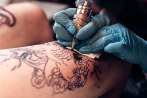 Nahaufnahme eines Tätowierers, der blaue Handschuhe trägt und mit einer Tätowiermaschine ein detailliertes Motiv auf die Haut einer Person tätowiert.