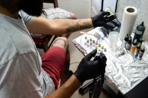 Eine tätowierte Person lässt sich eine Tätowierung auf den Arm stechen. Tätowierer mit Handschuhen, umgeben von Tintenflaschen und Ausrüstung auf einem mit Folie bedeckten Tisch.