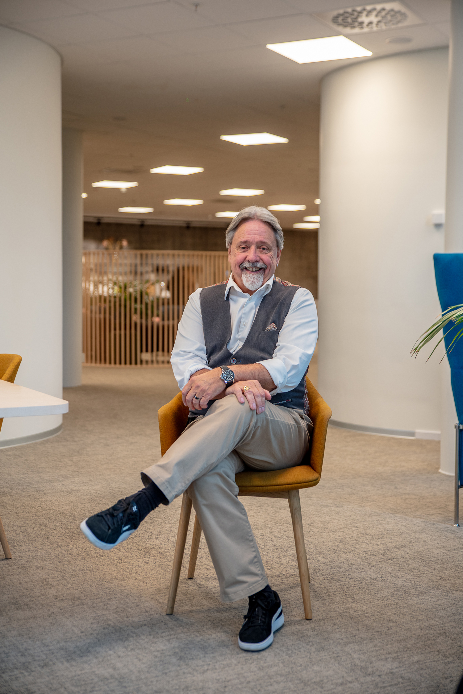 Smiling man with a beard and gray hair, seated in a modern office area, wearing a vest, seated on a mustard chair, with one leg crossed.