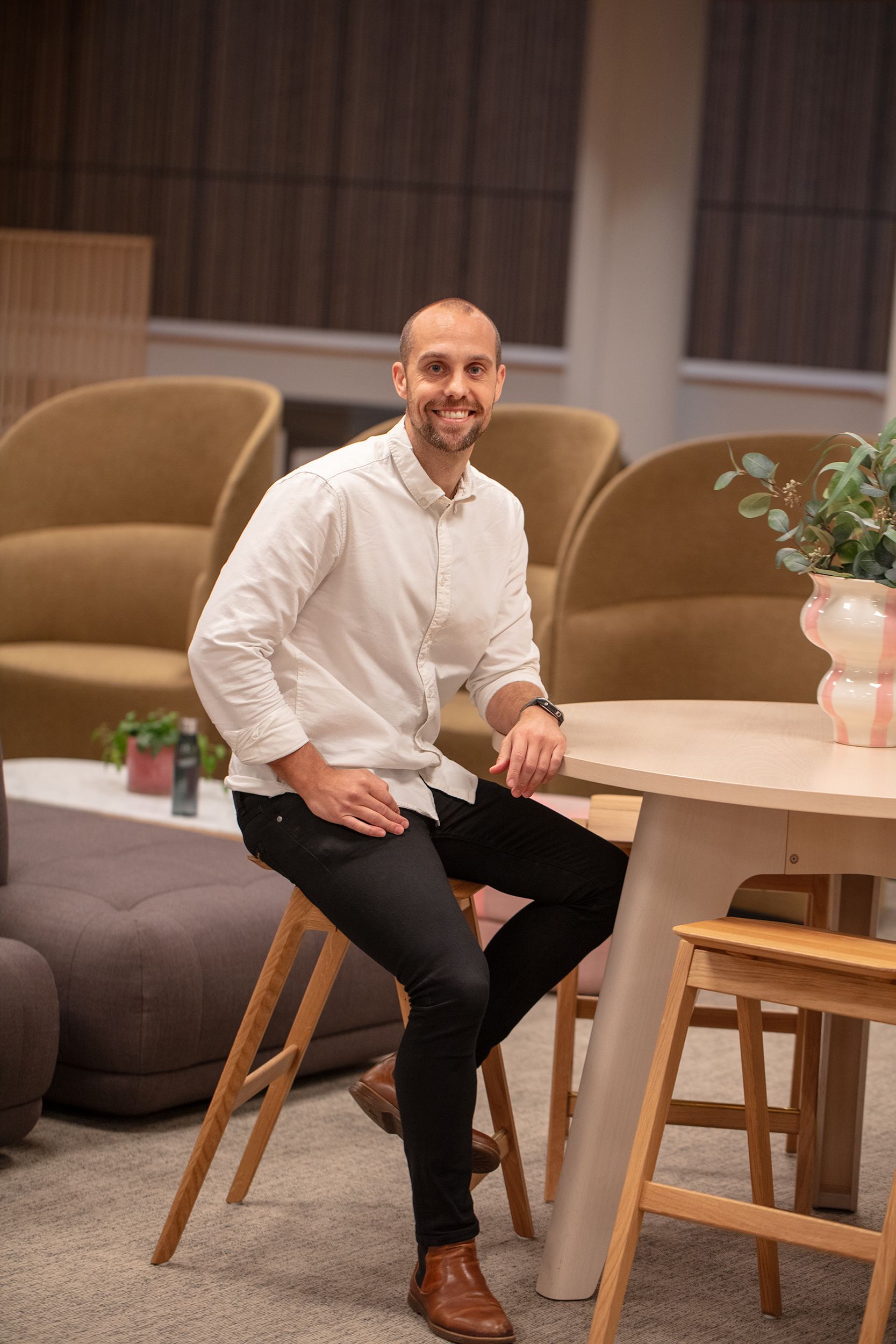 Man in a white shirt sitting on a wooden stool by a round table, smiling in a cozy, modern lounge area.