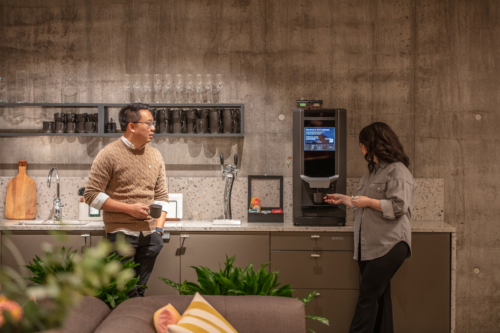 Two people in a modern kitchen area; one using a coffee machine, the other holding a mug. Shelves with glasses and mugs are in the background.