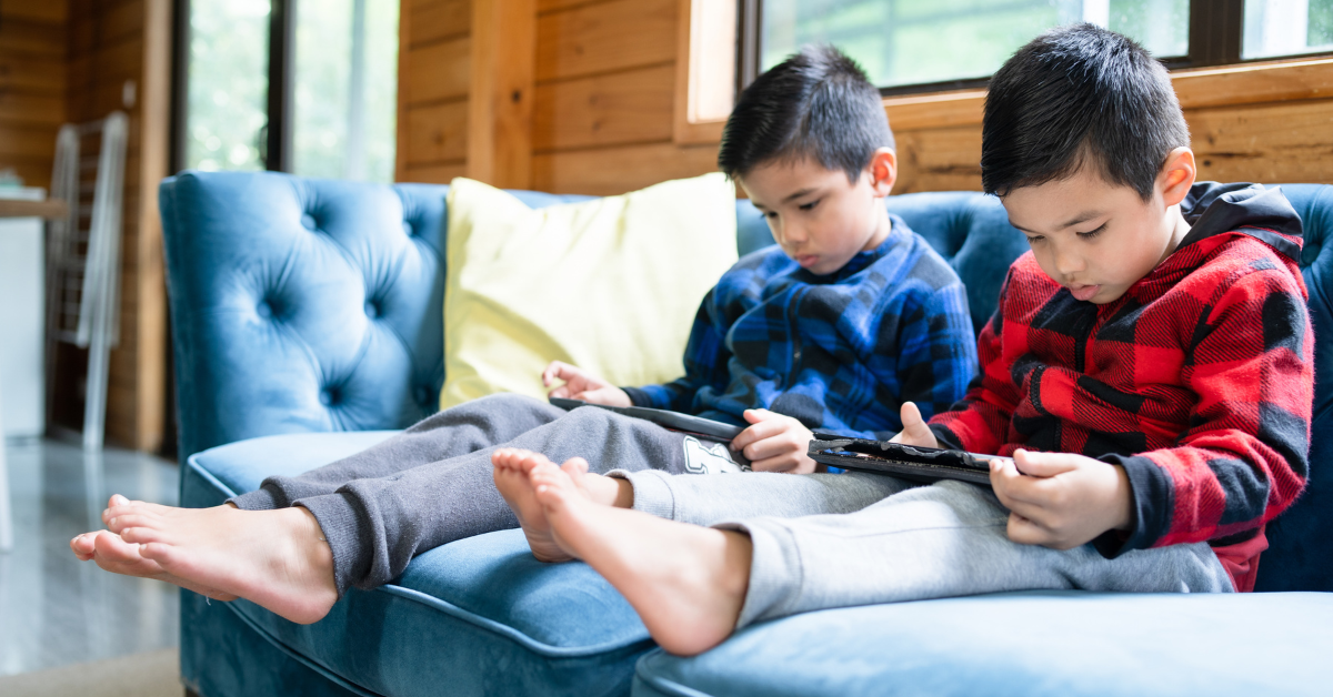 Two little boys sitting on a couch, both staring at their tablets