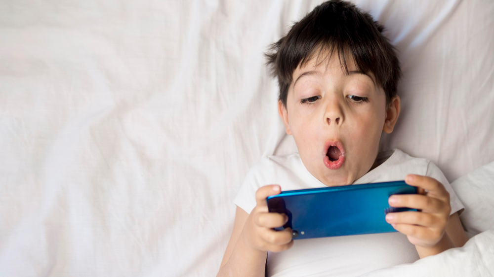 Boy in bed, looking at a blue smartphone and making a surprised and shocked face