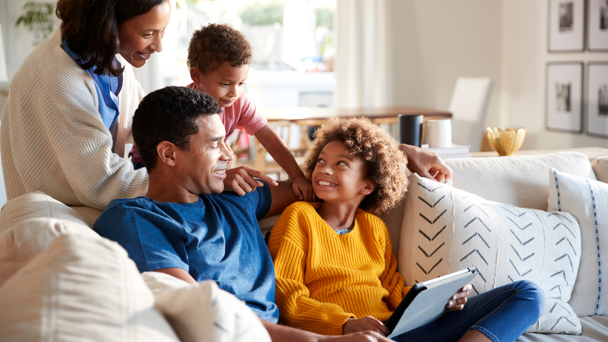 Dad and daughter on couch, daughter looks up to her mother and brother, who are standing behind the couch, all are smiling