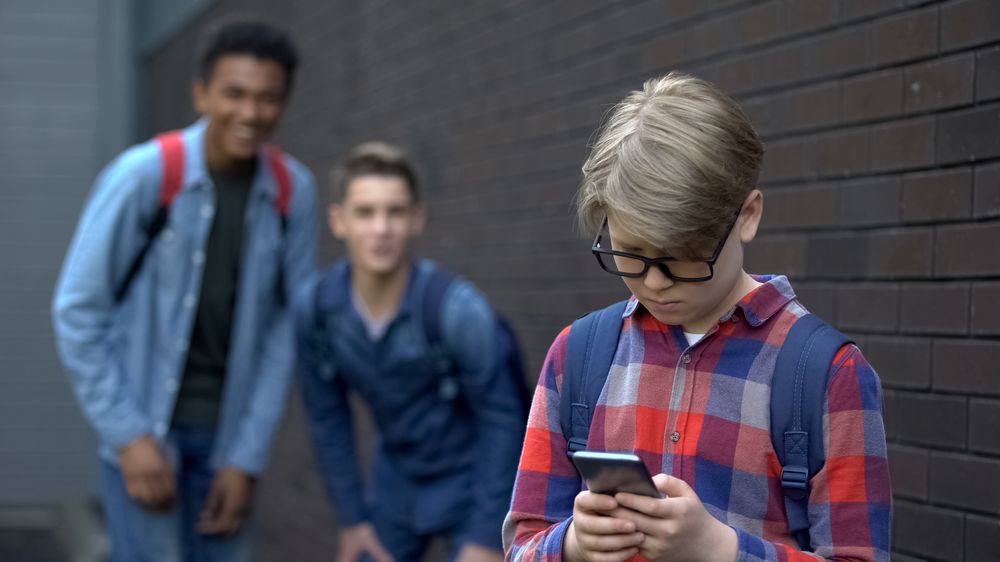 Ein Junge schaut traurig auf sein Handy, hinter ihm stehen zwei Mitschüler, die ihn auslachen, Cybermobbing