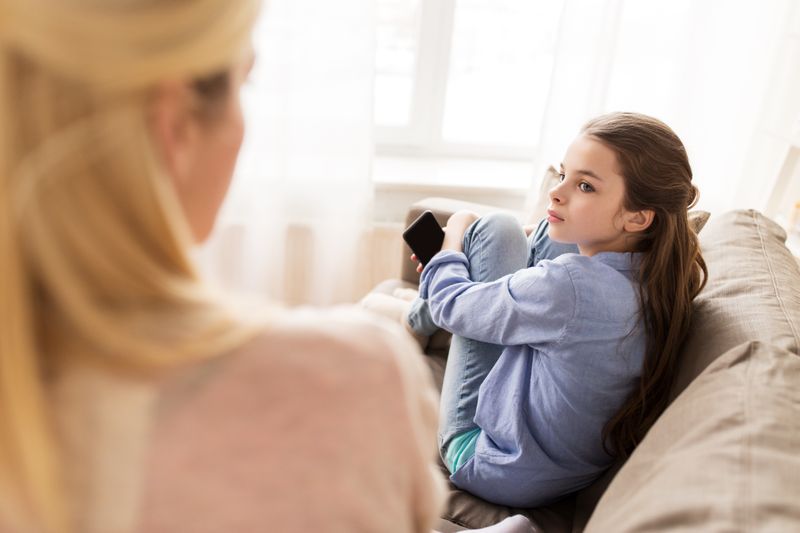 Trauriges Mädchen sieht auf der Couch, hat Handy in der Hand und sieht Mutter an