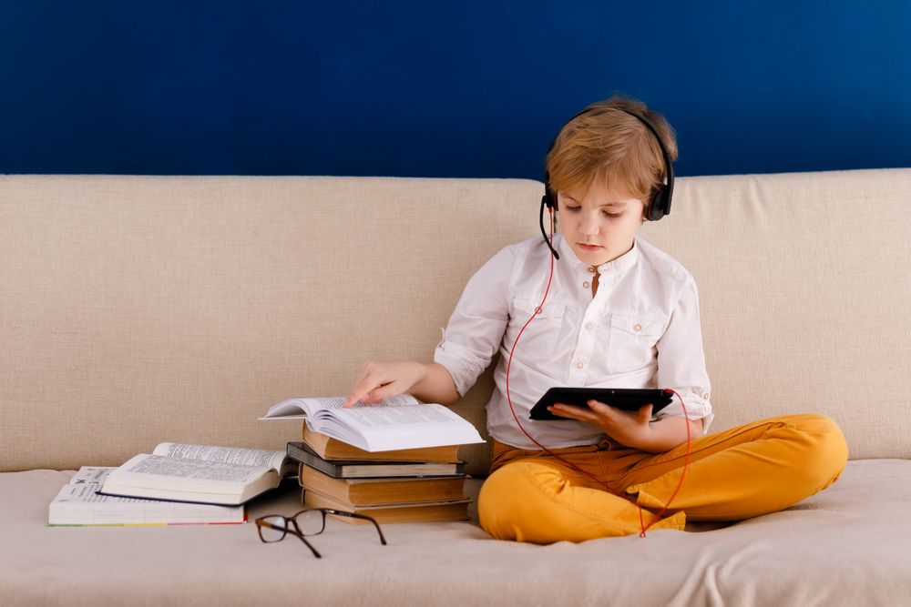 Junge sitzt auf der Couch und macht mit seinem Tablet Hausaufgaben, neben ihm liegen Lernbücher und eine Brille