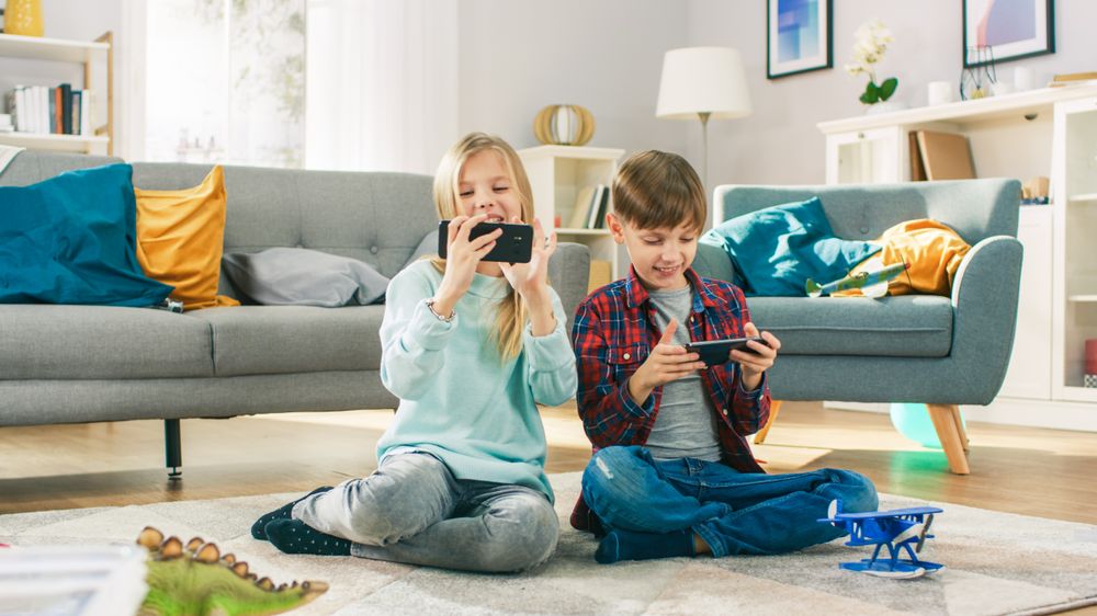 Ein Mädchen und ein Junge sitzen am Wohnzimmerboden und spielen auf ihren Handys