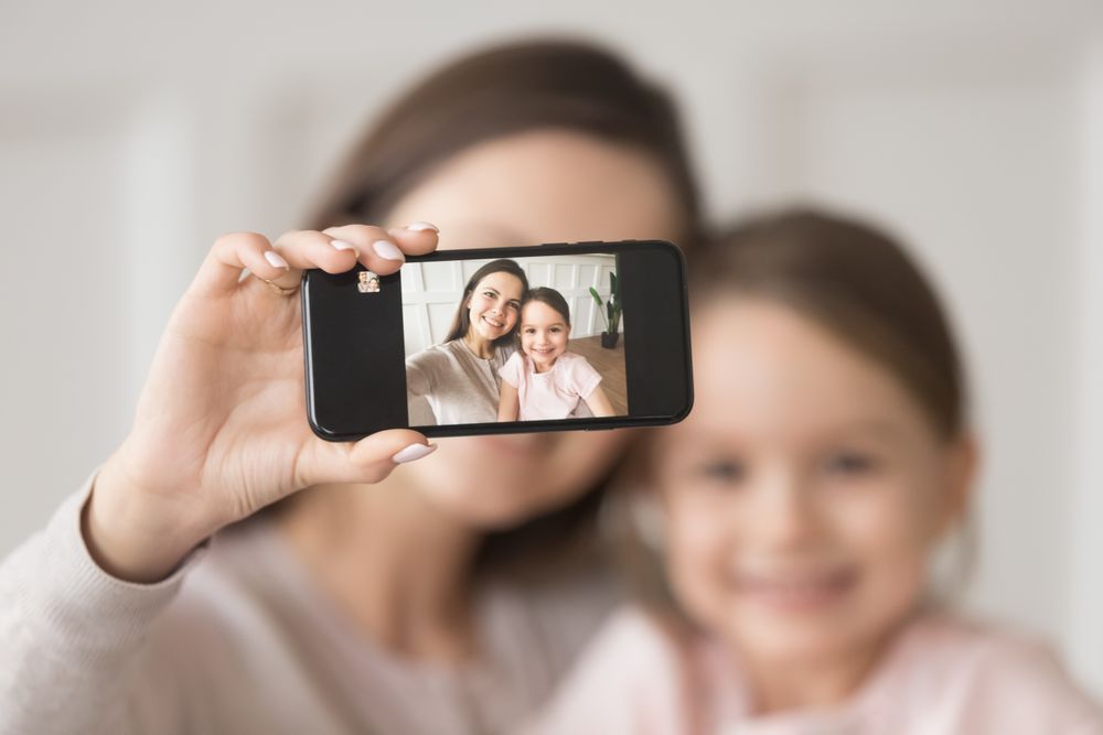 Glückliche junge Mutter hält Handy, macht Selfie auf Handy, umarmt Kind Tochter, lächelnd Mutter und Kind Mädchen Blick auf Smartphone-Kamera machen Foto zusammen, Fokus auf Handy-Bildschirm Porträt