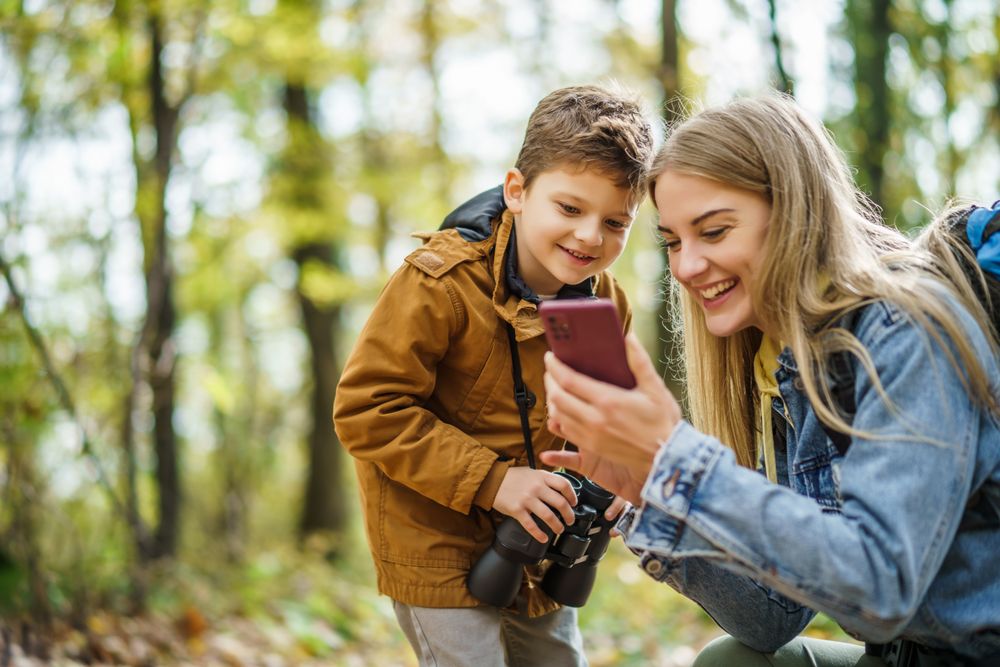 Mama und Sohn im Wald, sie lächeln während sie gemeinsam auf ein Handy schauen