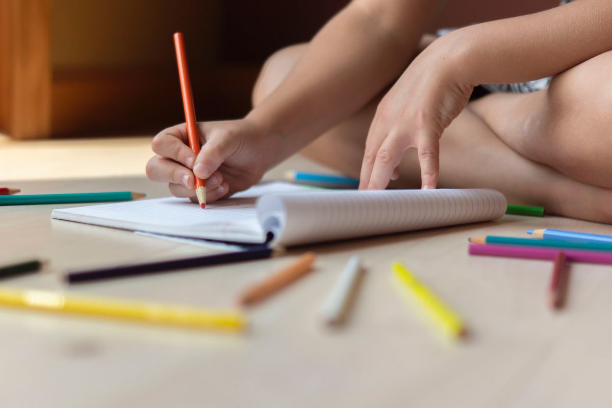 Child is sitting on floor, drawing in a notebook with colorful pencils