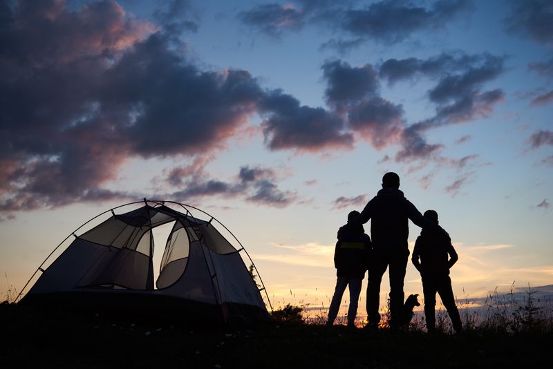 Vater mit zwei Kindern und Hund stehen neben einem Zelt und beobachten den Sonnenuntergang