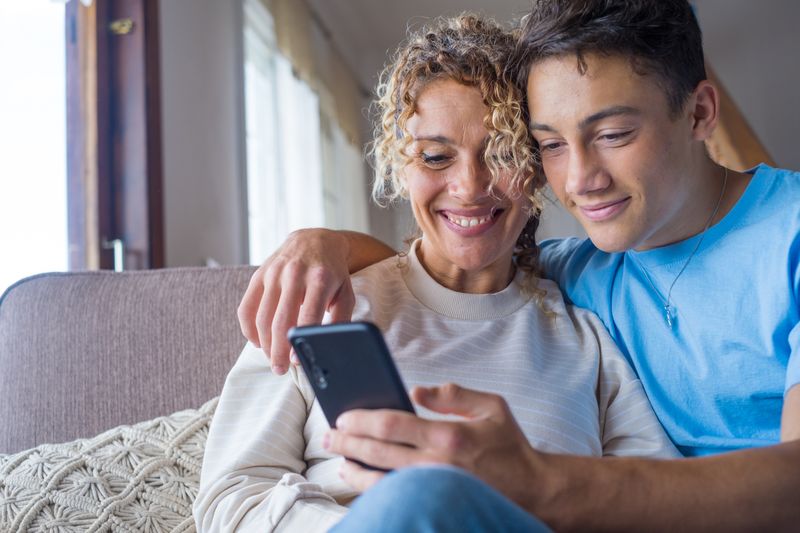 Jugendlicher sitzt mit seiner Mutter auf der Couch, sie schauen gemeinsam lächelnd auf ein Handy