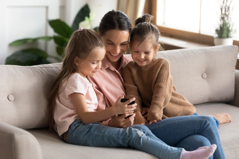 Mutter sitzt mit ihren jungen Töchtern auf dem Sofa, sie lächeln alle und schauen gemeinsam auf ein Handy