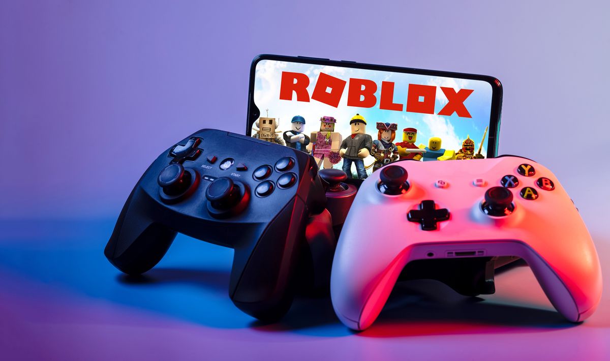 Two video console controllers, phone in the background showing start screen of Roblox