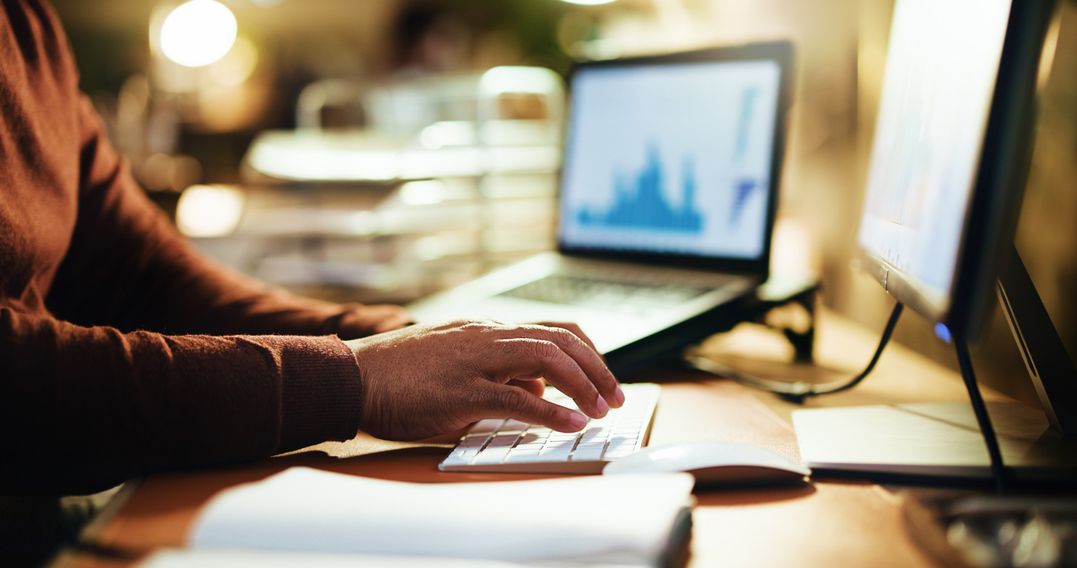 Person typing on a keyboard at a desk with a monitor and laptop displaying graphs in a softly lit workspace.